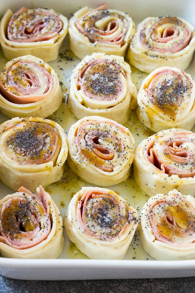 Ham and Cheese Pinwheels Recipe | Ham and Cheese Roll Ups | Ham and Cheese Rolls | Ham and Cheese Bread Rolls #ham #cheese #appetizer #pinwheels #uzhal Dough rolled up with ham and cheese and topped with a poppy seed butter in a ceramic dish.