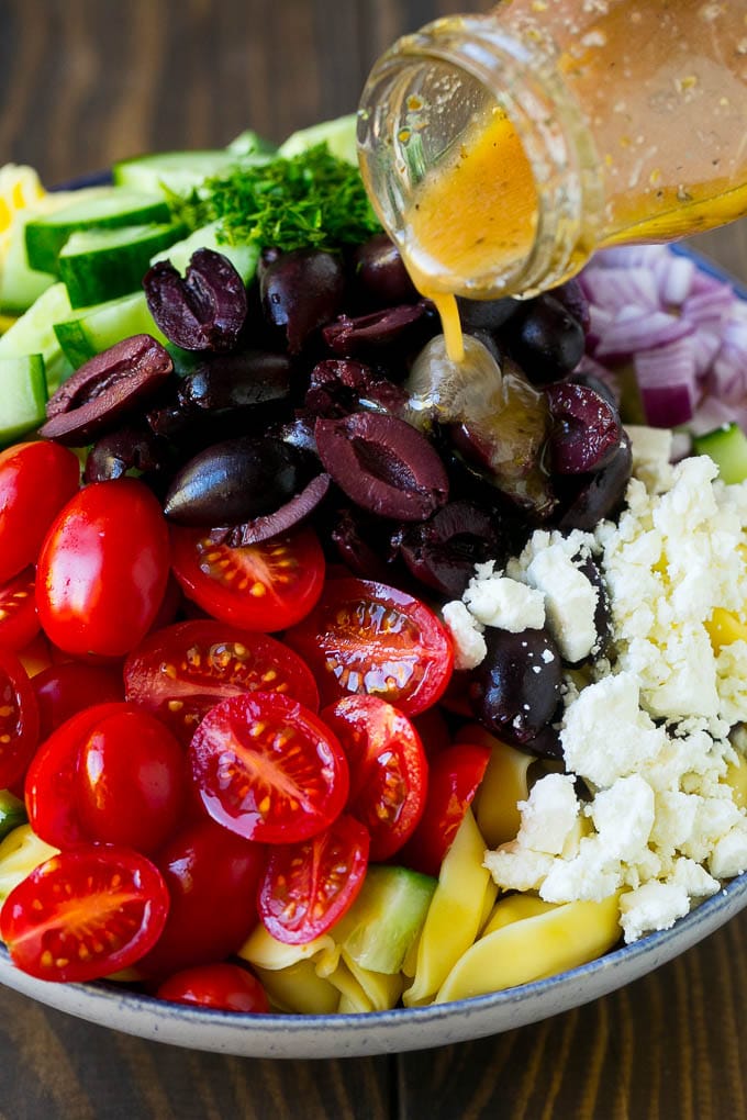Greek Tortellini Salad Recipe | Pasta Salad | Tortellini Salad | Greek Salad #greek #tortellini #pasta #salad #cucumbers #olives #dinner #uzhal Salad dressing being poured over a bowl of tortellini and vegetables.