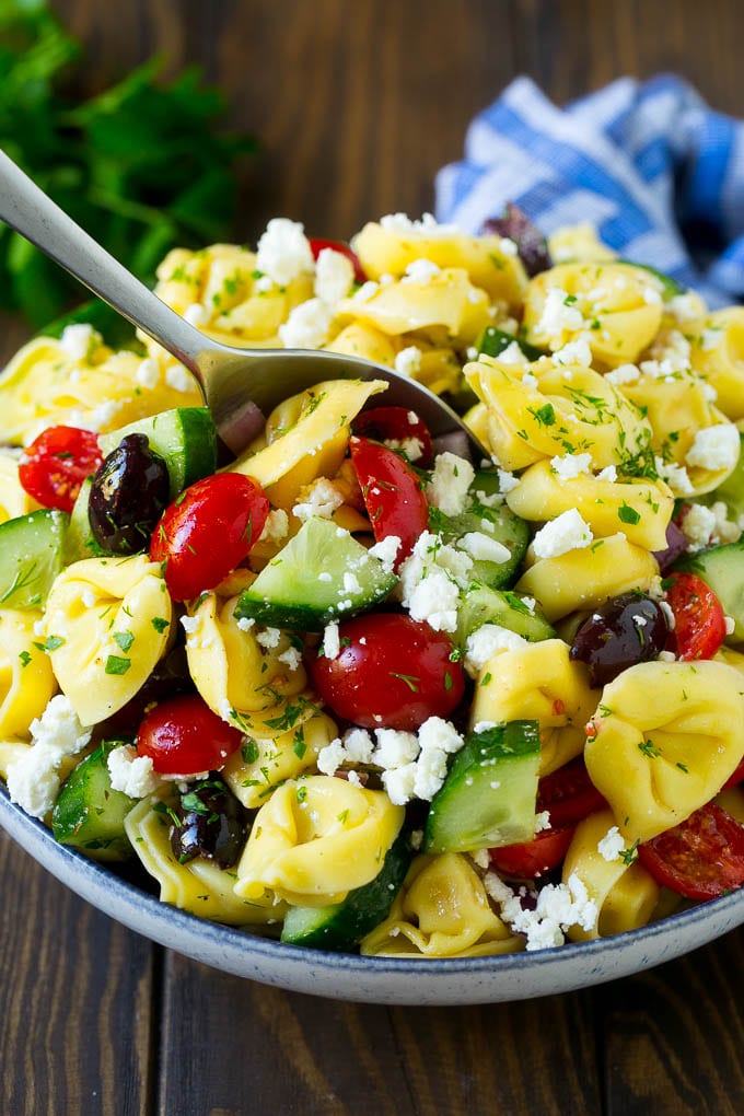 Greek Tortellini Salad Recipe | Pasta Salad | Tortellini Salad | Greek Salad #greek #tortellini #pasta #salad #cucumbers #olives #dinner #uzhal A bowl of Greek tortellini salad with a serving spoon in it.