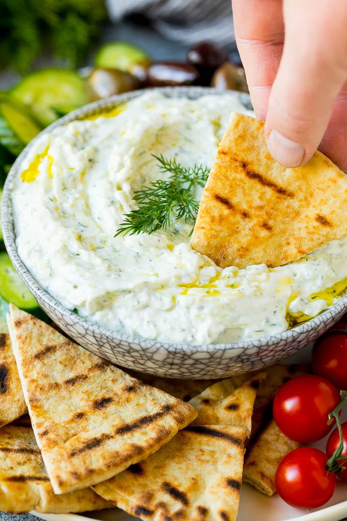 The best homemade tzatziki sauce with cucumber and herbs mixed into Greek yogurt. #appetizer #uzhal A hand dipping pita bread into a bowl of tzatziki.