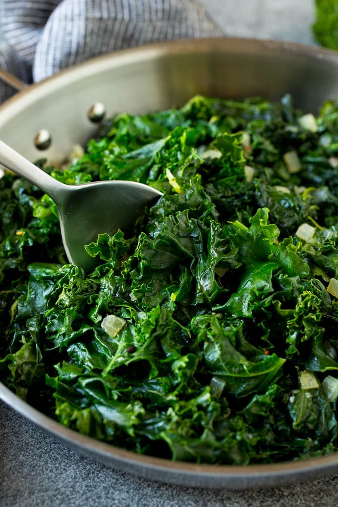 Sauteed kale flavored with olive oil and garlic, a quick and easy side dish! #kale #uzhal A pan of sauteed kale with a serving spoon in it.