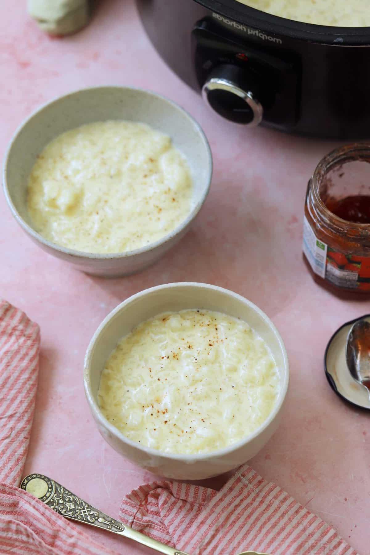 Due ciotole di budino di riso in slow cooker davanti a una crock pot e un barattolo aperto di marmellata.