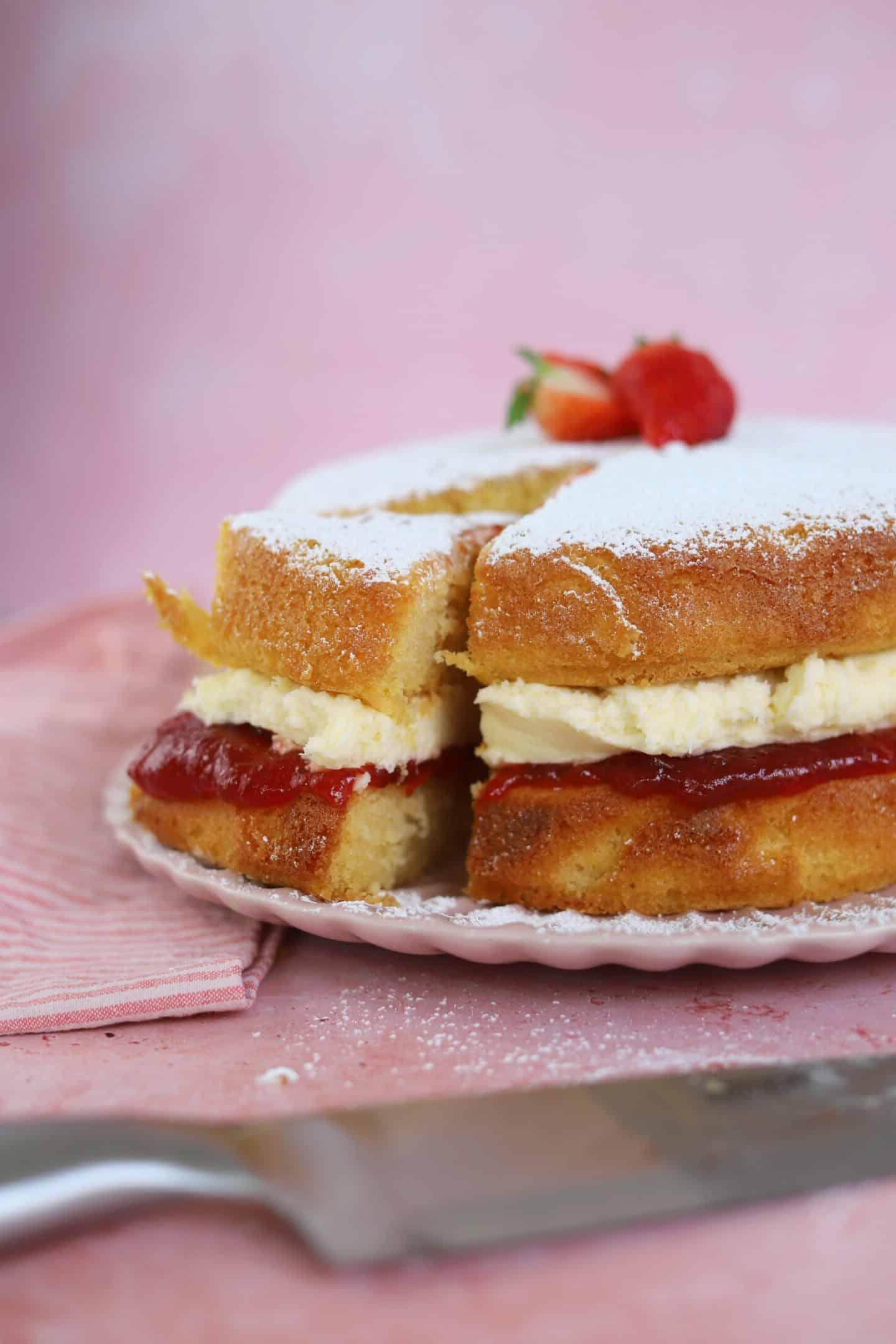 Torta Victoria Senza Glutine 3 Una deliziosa Torta Victoria senza glutine, con una fetta già tagliata, che rivela i suoi strati di pan di Spagna, burrocrema e marmellata, su un piatto elegante.