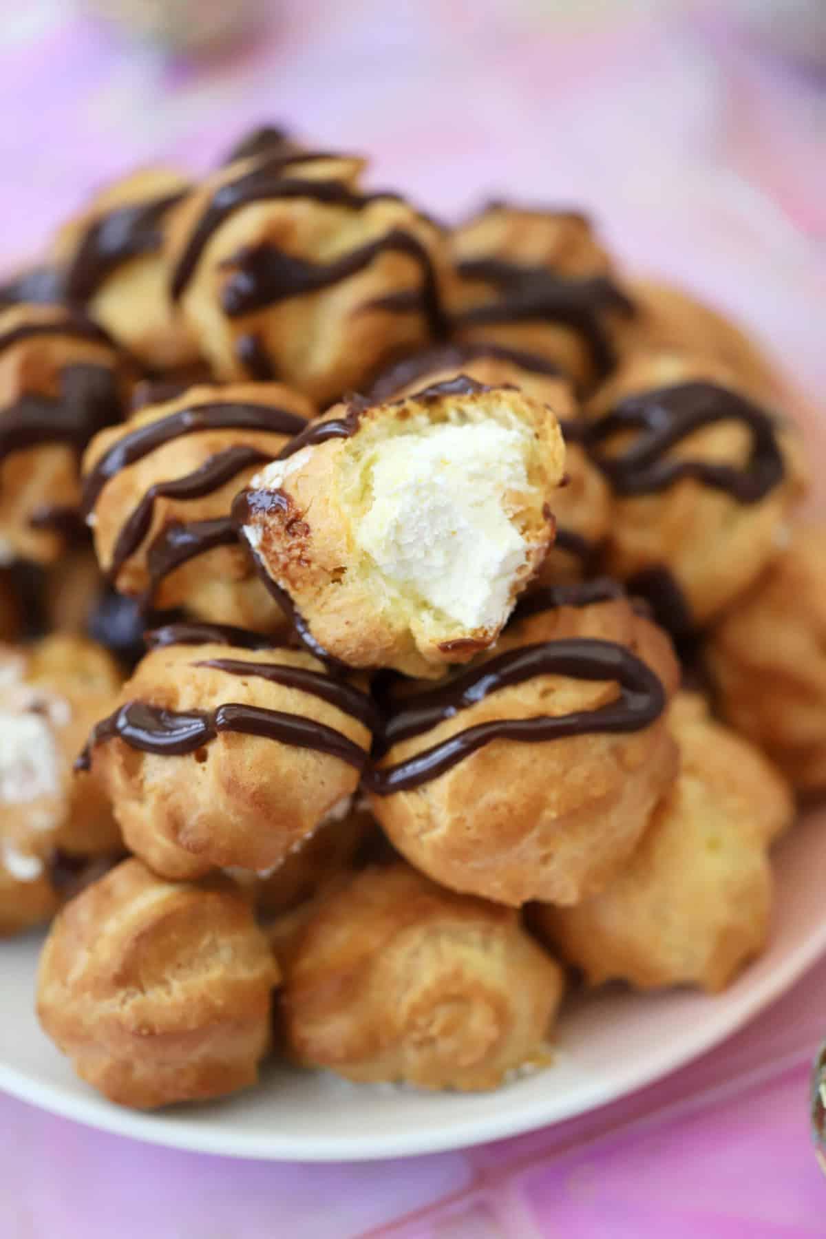 25+ Dolci Natalizi Senza Glutine 26 A plate of gluten free profiteroles drizzled with chocolate sauce, with one cut open to show the whipped cream filling.