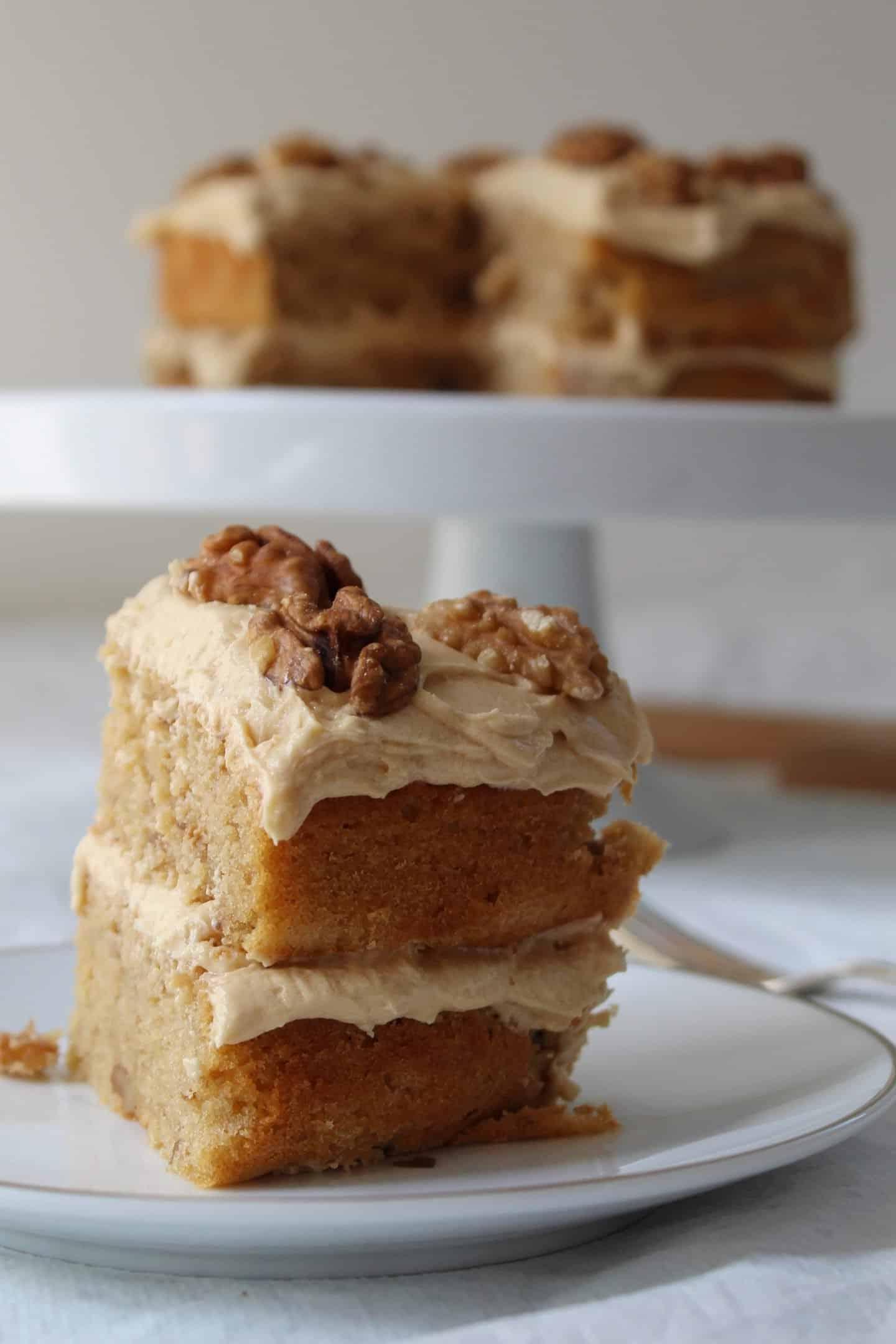 Torta al Caffè e Noci Senza Glutine 5 Una fetta di torta al caffè e noci senza glutine con l'intera torta su un'alzata dietro.