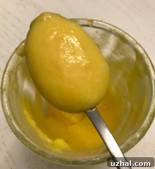 Homemade Lemon Curd in a clear glass jar, with fresh lemon slices and a spoon, illustrating its smooth texture and vibrant yellow color.