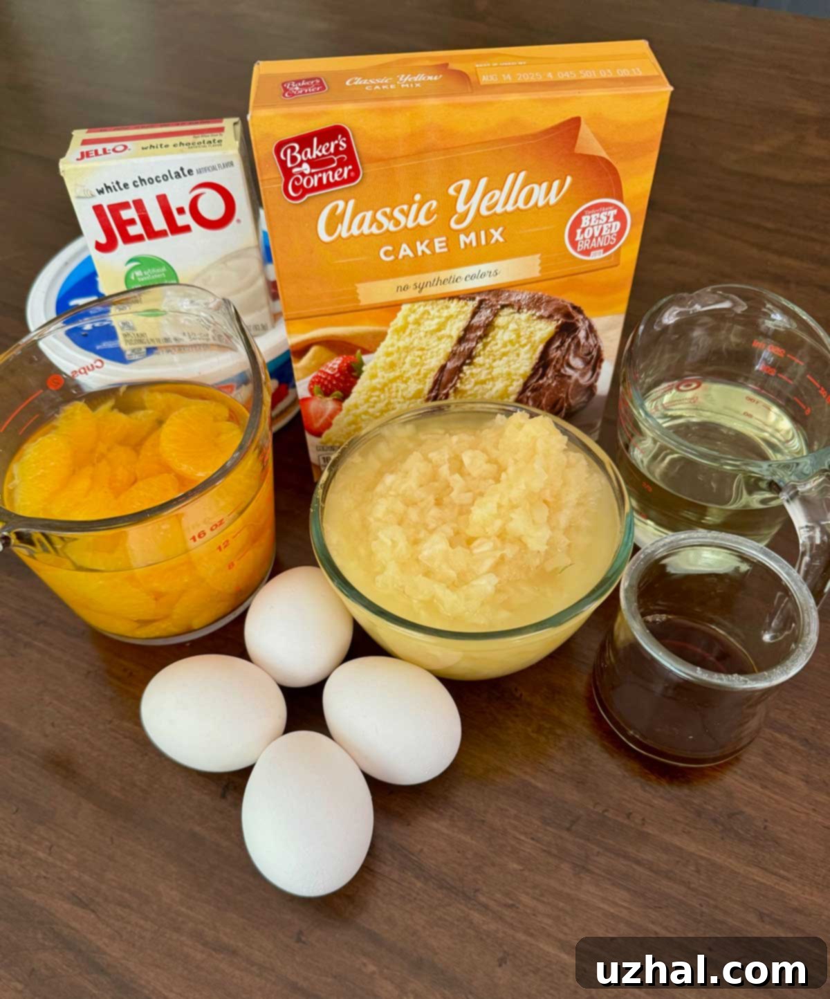 Ingredients for Pig Pickin' Cake including pudding mix, yellow cake mix, mandarin oranges, eggs, oil, pineapple, vanilla and whipped topping, laid out on a kitchen counter.