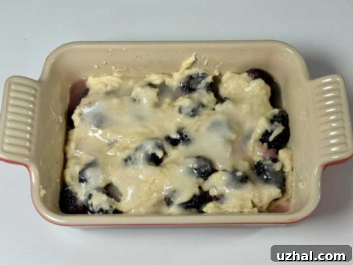 Boiling water being carefully poured over the sugar and cornstarch layer of a blackberry cobbler, creating a glossy surface.